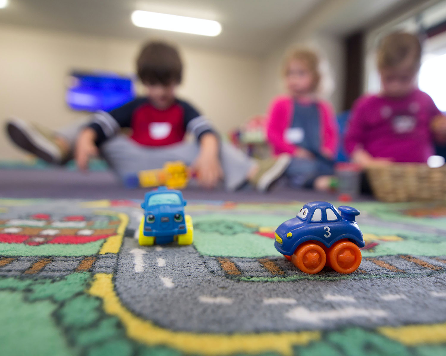 Toy cars on a play mat with kids in the background.