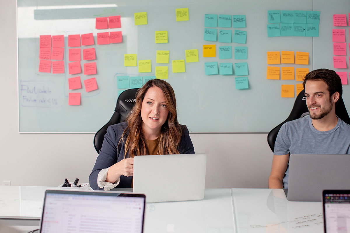 Coworkers sit at a table working on laptops with bright colored sticky notes behind them.