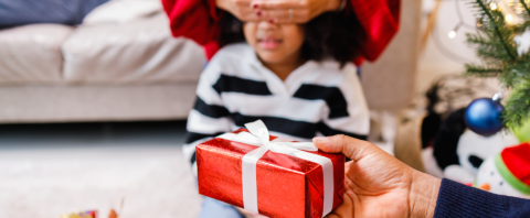 A young girl waits to open a holiday gift under a Christmas tree.